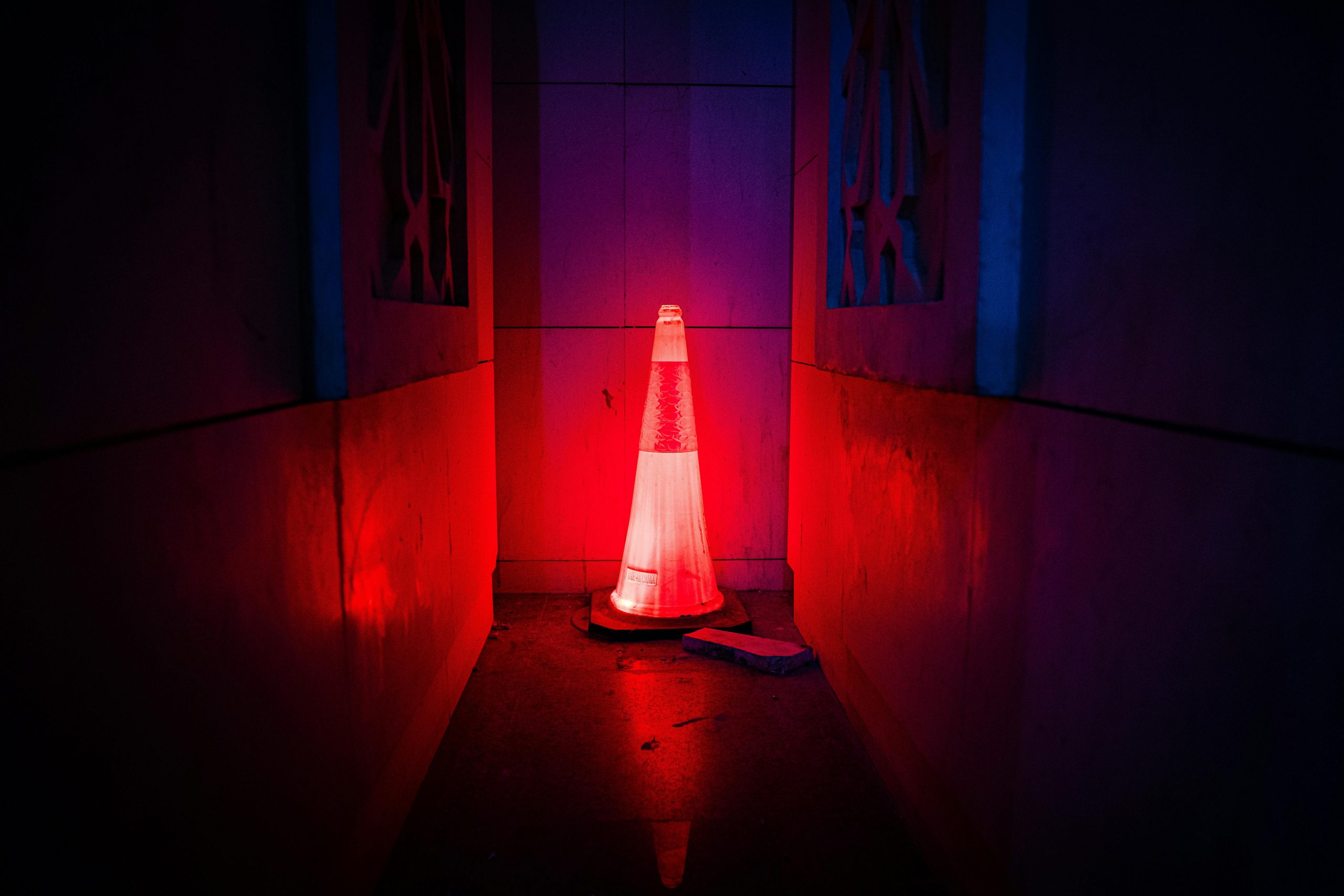 A red neon-lit traffic cone emits a mysterious glow in a dark urban alleyway, creating an enigmatic atmosphere.